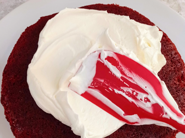 una espátula untando glaseado de Philadelphia encima de una capa de pastel red velvet